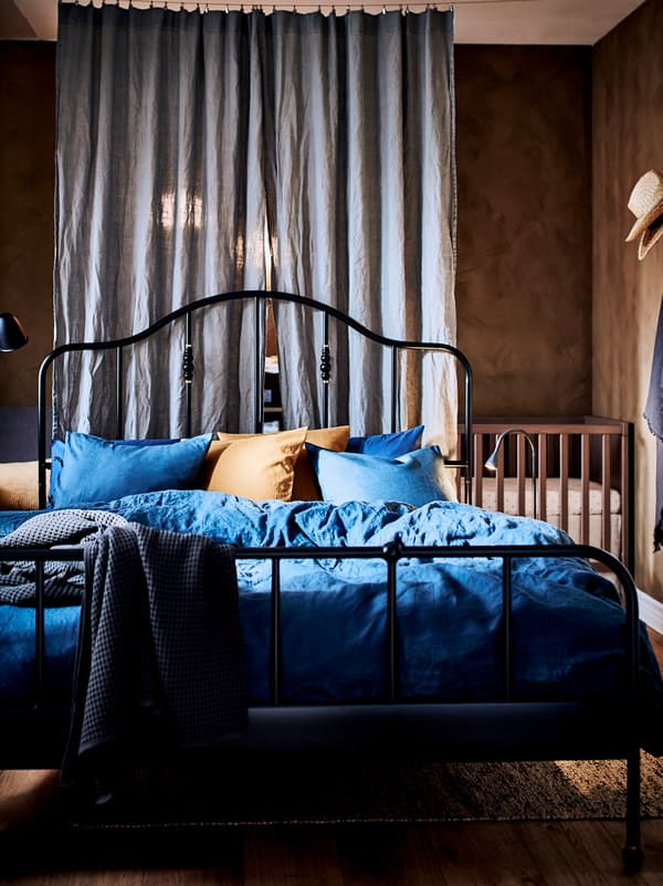 Bedroom with a black SAGSTUA bed and, in a corner behind a room-dividing curtain, stands a wooden baby cot.