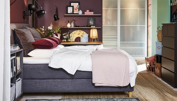 Bedroom with a dark grey divan bed, black wall lamps, a jute rug, a black-brown chest of drawers and dark red cushion covers.