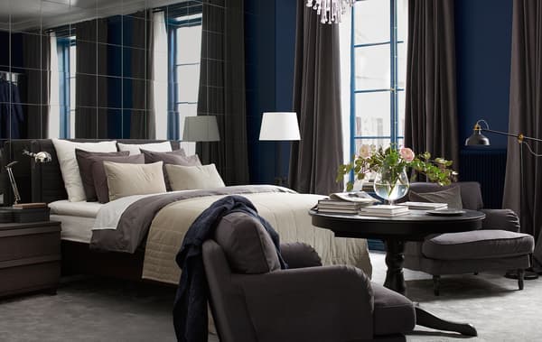 Bedroom with a wall of mirrors, a bed with lots of textiles, an armchair, floor lamps and fresh flowers.