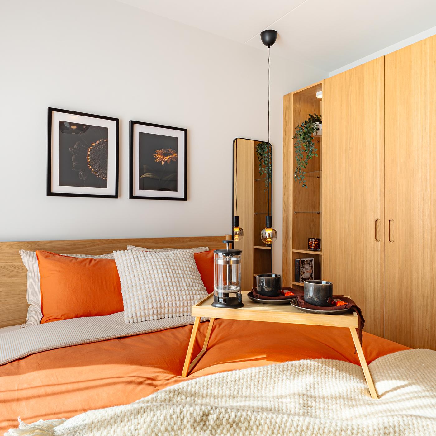 Bedroom with an oak veneer bed frame dressed in terracotta and neutral bedding, a breakfast tray on the bed, framed wall art above the headboard, pendant lights on both sides, and a large oak veneer wardrobe with a reeded glass door.