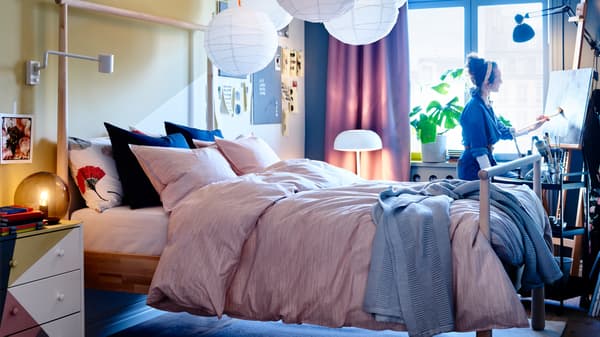Bedroom with GJÖRA bed frame in birch, white pendant lamps, pink bed textiles, a grey table lamp and lilac curtains.