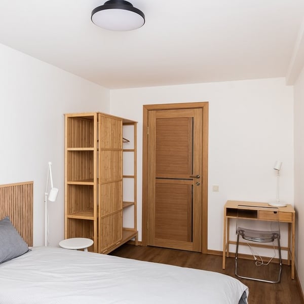 Bedroom with minimalist wooden furniture and desk – Modern bedroom interior featuring a wooden door, open wardrobe, small writing desk with lamp, and neatly made bed in neutral tones.