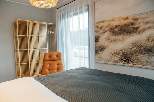 Bedroom with natural wood wardrobe and leather accent chair – Light-filled bedroom with a wooden open wardrobe, tan leather chair, and large wall art of coastal sand dunes.