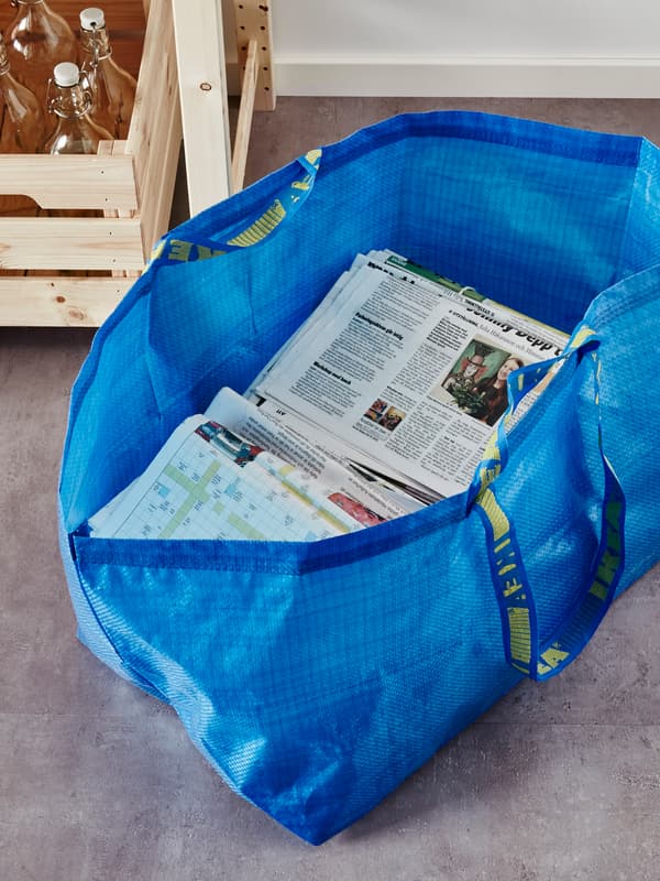 Beg FRAKTA berwarna biru, separuh penuh dengan surat khabar, di atas lantai di hadapan sekotak botol KORKEN kaca jelas dengan penyumbat.