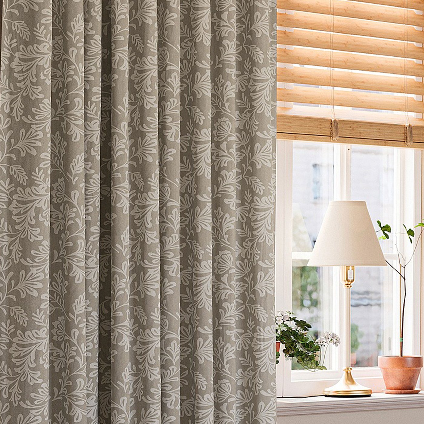 Beige gardin med subtilt blommönster bredvid ett fönster med träpersienner, en vit bordslampa och en liten krukväxt på fönsterbrädan.