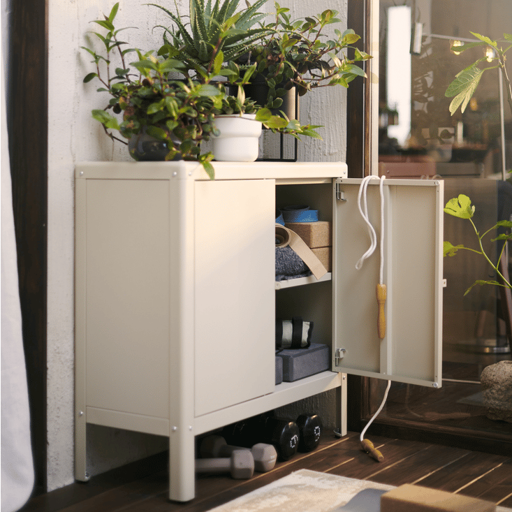 Beige KOLBJÖRN outdoor cabinet in blacony area with one door opened, showing pairs of shoes stored neatly in the cabinet with other items. Several plant pots are also displayed on top of the cabinet. 