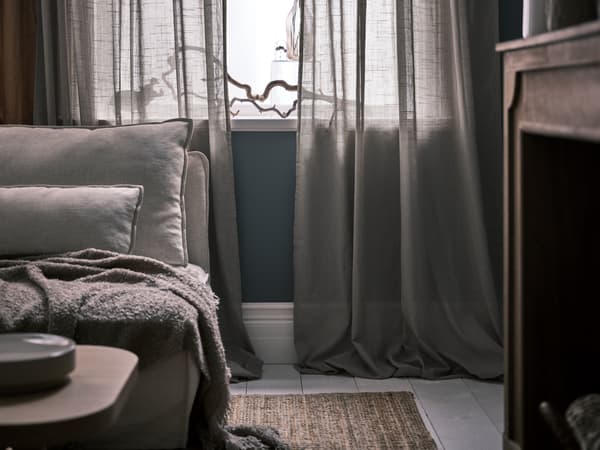 Beige SILVERLÖNN sheer curtains draped onto the floor in front of a window beside a sofa on a LOHALS jute rug.