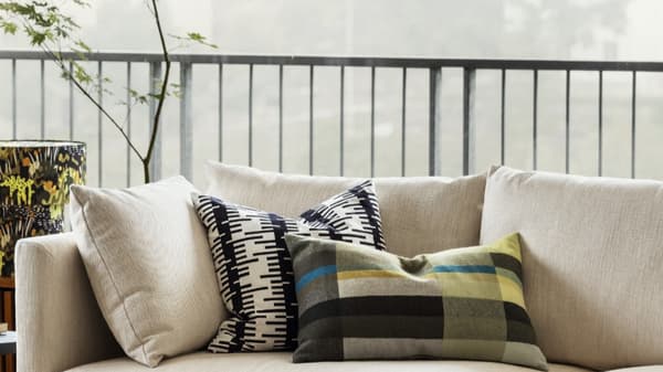 Beige soffa med flera dekorativa kuddar i olika mönster och färger, inklusive en svartvit geometrisk kudde och en grönrutig kudde med blå och gula detaljer. I bakgrunden syns ett räcke och suddig utsikt genom ett stort fönster.