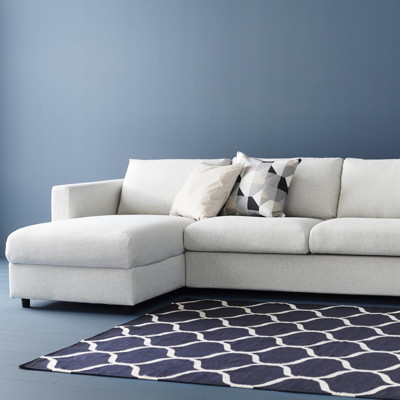 Beige VIMLE slaapbank met chaise longue tegen een blauwe muur, met blauw vloerkleed met motief. Op de zitbank liggen twee kussens.
