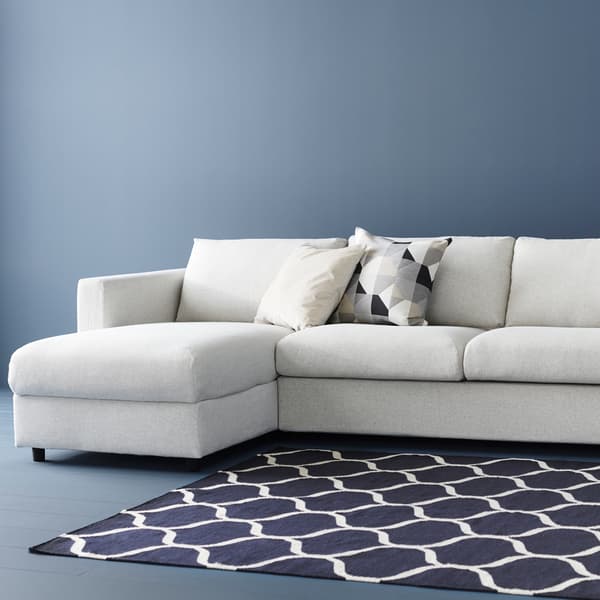 Beige VIMLE sofa bed with chaise longue against a blue wall, with blue patterned rug. There are two cushions on the sofa.