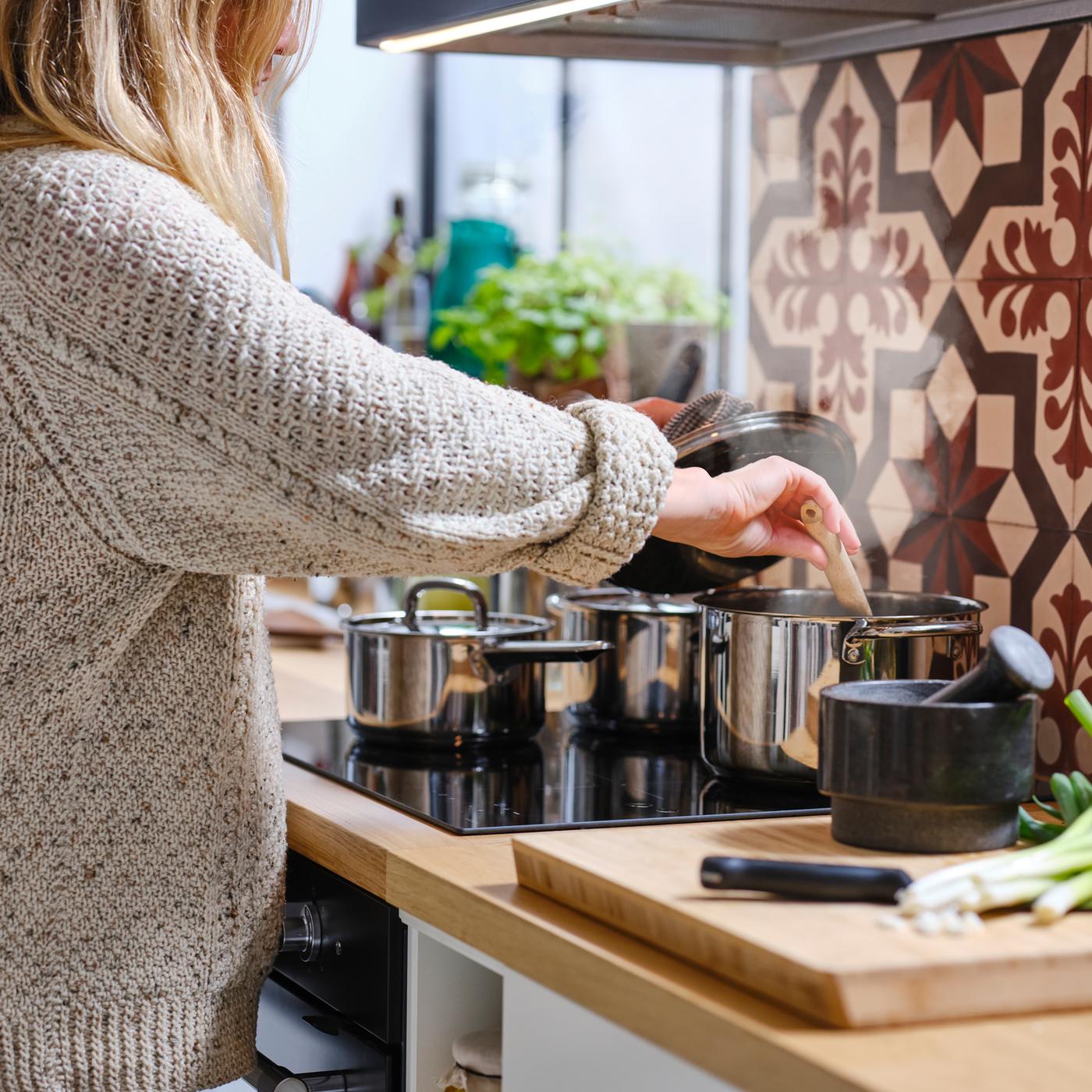 Beigeen neuleeseen pukeutunut henkilö nostaa induktiokeittotasolla olevan ruostumattomasta teräksestä valmistetun IKEA 365+ -kattilan kantta ja sekoittaa kattilan sisältöä.