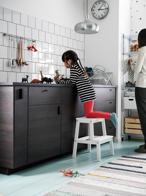 เด็กผู้หญิงคุกเข่าอยู่บนเก้าอี้เตี้ย BEKVÄM/เบียแควม เล่นตุ๊กตารูปสัตว์ตัวเล็กอยู่บนท็อปครัวสีเข้ม มีผู้หญิงยืนอยู่ใกล้ ๆ