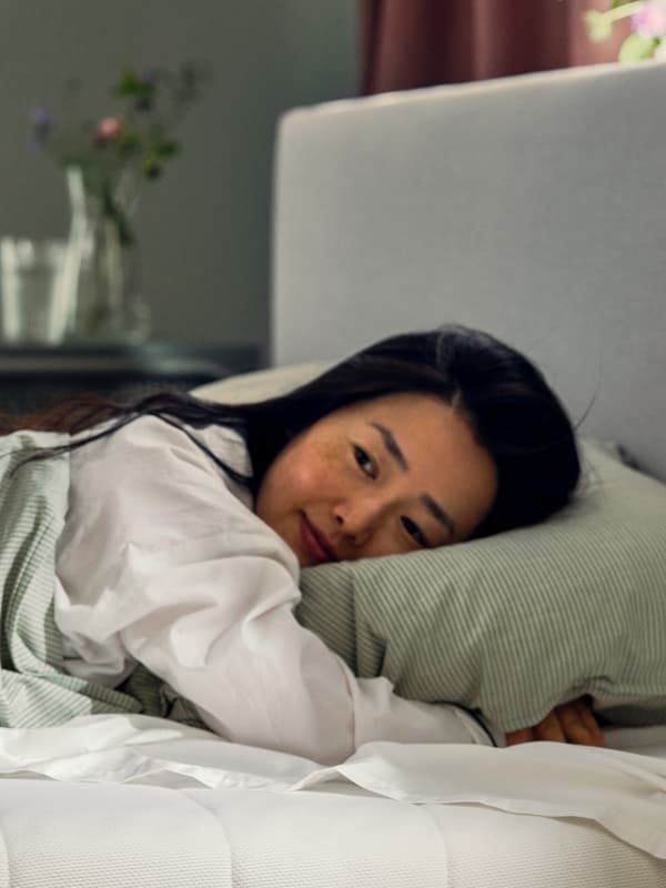 웃는 사람이 침대에 엎드려 누워 있습니다. 그들은 BERGPALM 베갯잇과 이불 커버에 낮은 베개를 껴안고 있습니다.