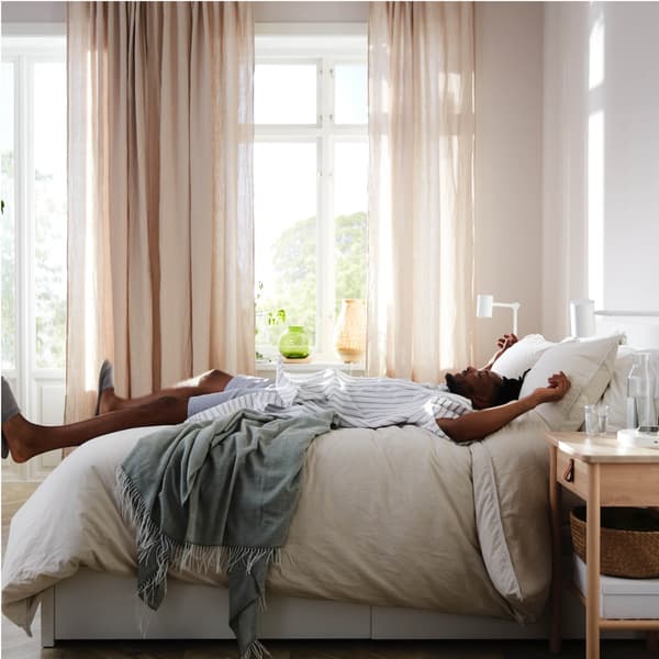 Berken BJÖRKSNÄS bed met bedlinnen in wit en beige naast een wit nachtkastje, een tafellamp in bamboe en een luchtreiniger.