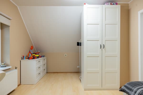Bērnu istabas uzglabāšanas un gulēšanas zona ar gultu, baltām atvilktnēm un augstu drēbju skapi pie slīpas sienas.