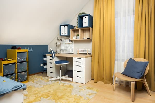 Bērnu mācību zona ar rakstāmgaldu, ergonomisku krēslu, sienas plauktiem un uzglabāšanas mēbelēm pie loga ar dzelteniem aizkariem.