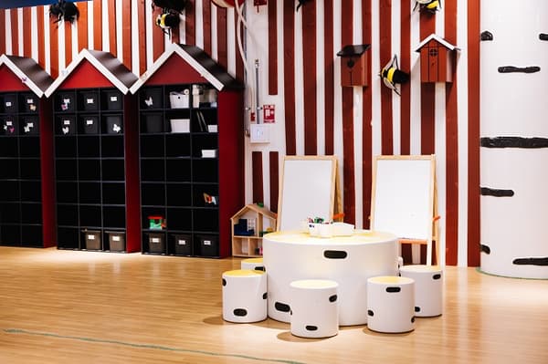 Bērnu radošā rotaļu zona ar apaļu galdu un baltiem krēsliem, kas atgādina kauliņus, ko ieskauj mākslas piederumi un molberti. Fonā ir sarkanbalti svītrainas sienas, melni, mazi plaukti māju formā, kā arī dekoratīvi putnu būrīši un koki.