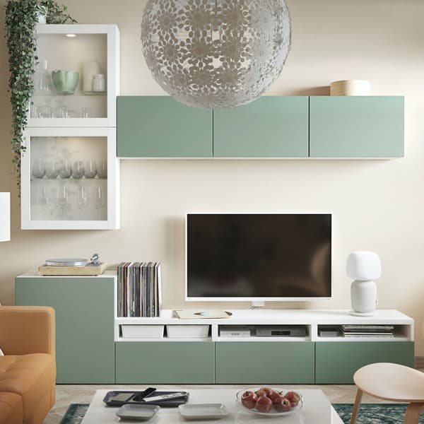 BESTÅ struttura bianca e verde in un salotto. Composizione con strutture ad anta in vetro e anta colorata. La TV è agganciata a parete e si intravede un divano, una poltrona e un tavolino. In altro si vede una lampada da soffitto.