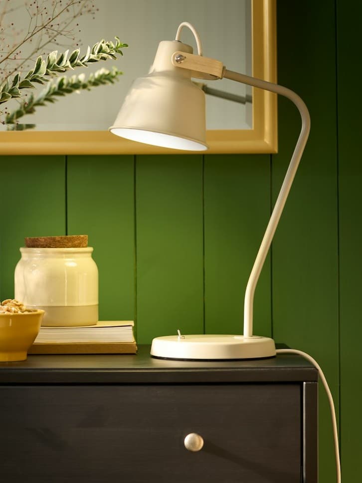 Beżowa lampa biurkowa stojąca na brązowej komodzie na tle zielonej ściany z lustrem.