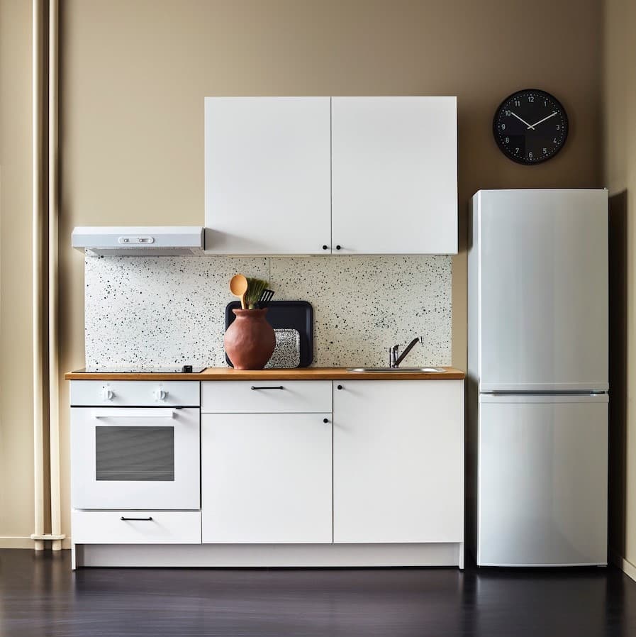 Biała kuchnia KNOXHULT wraz z białym AGD stoi na beżowej ścianie.