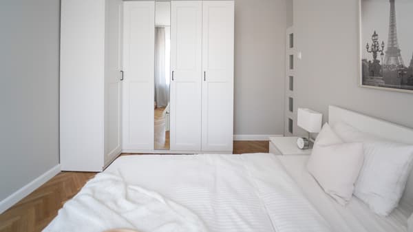 Biała sypialnia z białą, narożną sofą PAX i łóżkiem z białą pościelą.