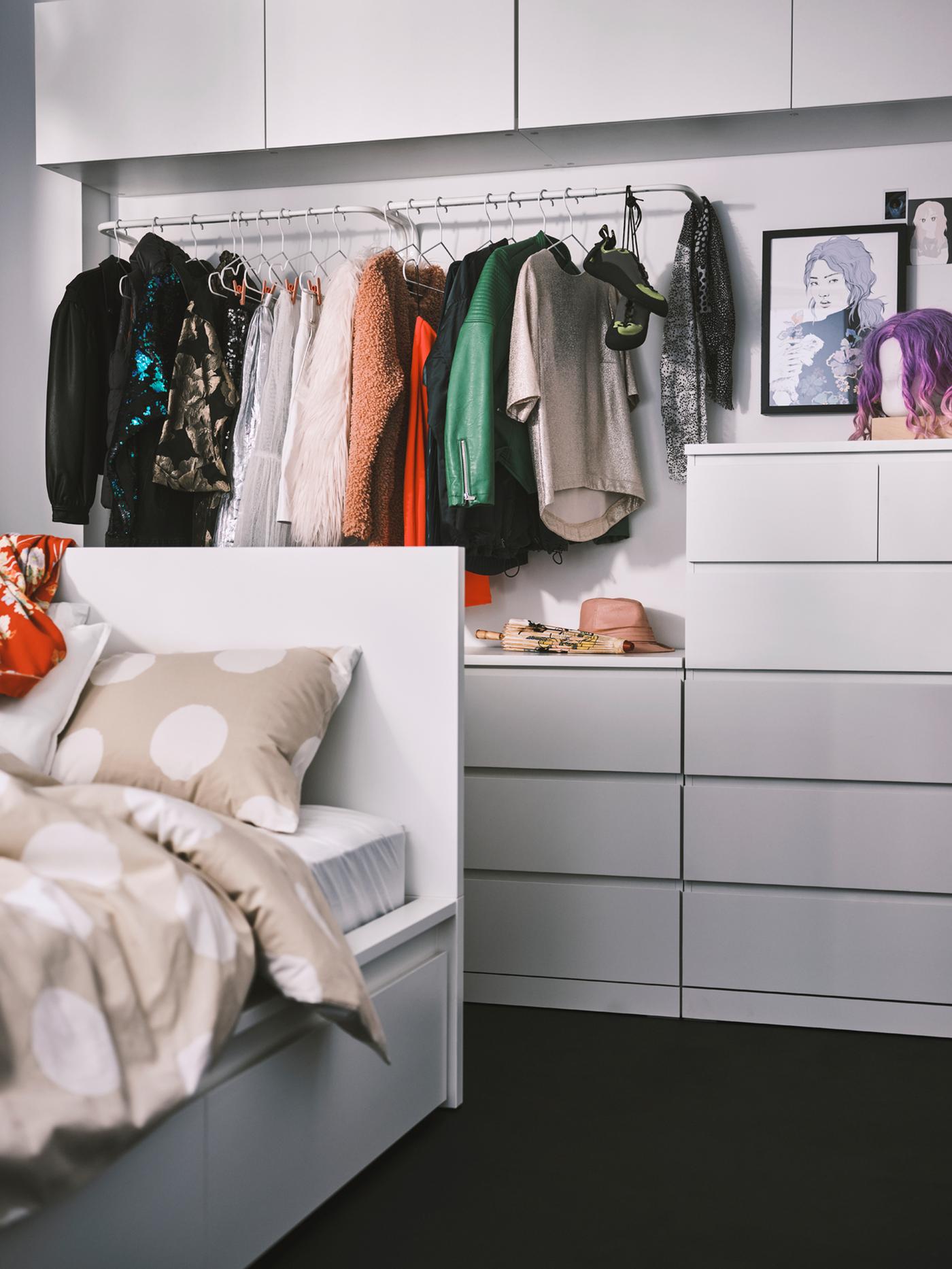 Białe łóżko Luröy/MALM z dwoma pojemnikami do przechowywania, a za nim, obok białej komody MALM z sześcioma szufladami, wiszą ubrania.  
