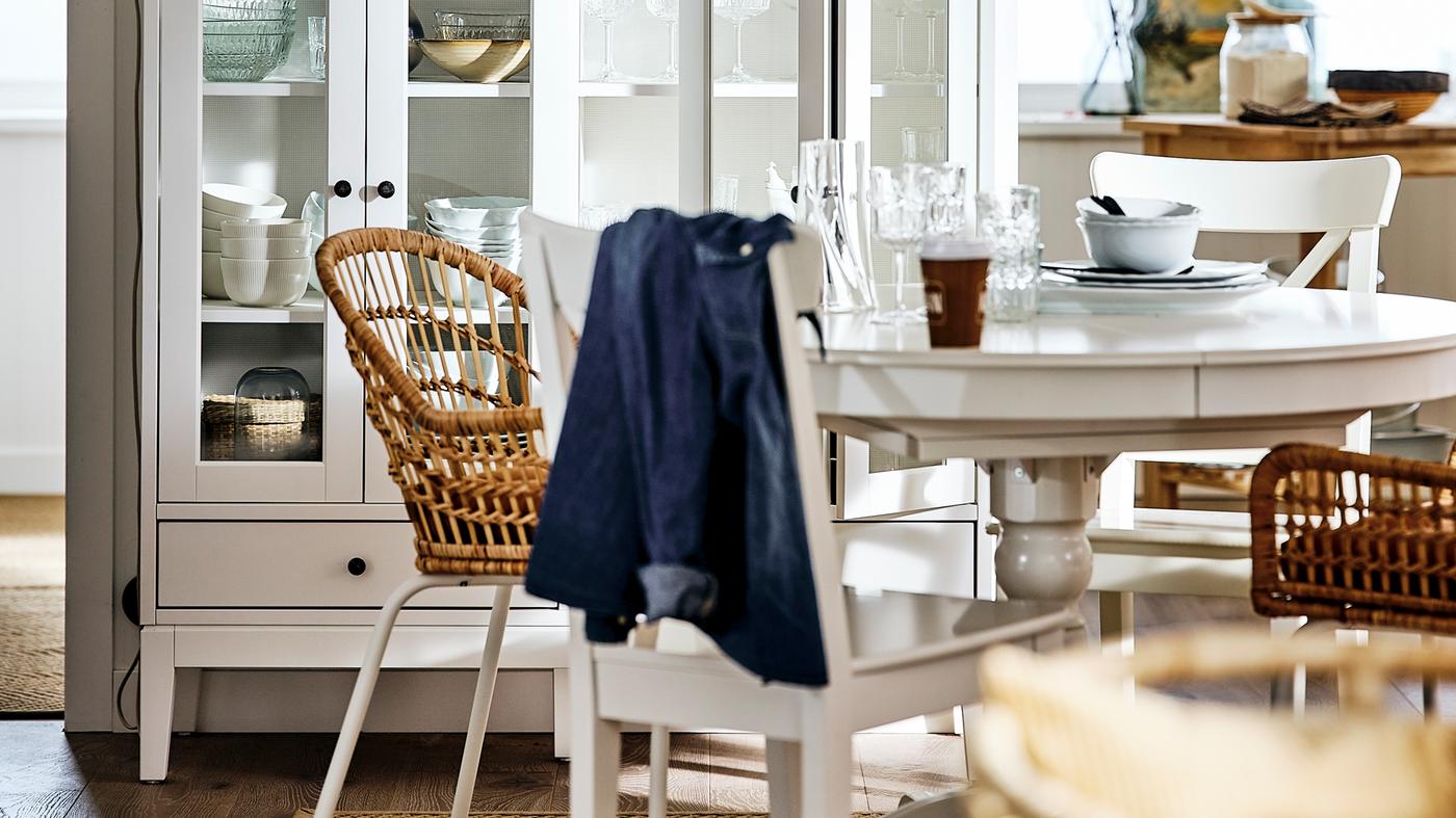 Biały rozkładany stół INGATORP w jadalni z talerzami i ze szklankami na blacie oraz z drewnianymi i rattanowymi krzesłami.