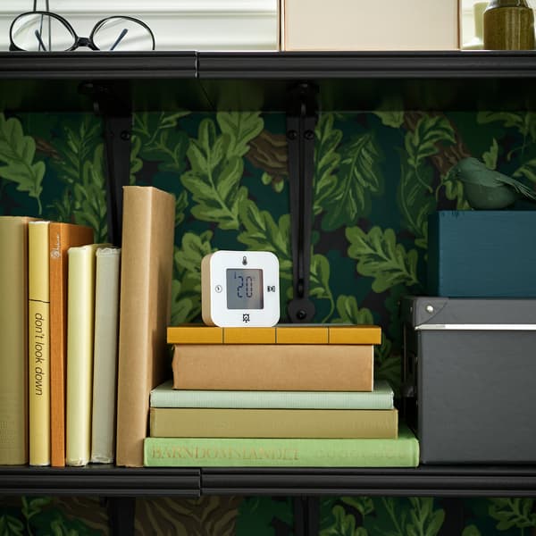 Biały zegar/termometr/budzik/minutnik KLOCKIS stojący na książkach na półce, pokazujący temperaturę w pomieszczeniu.