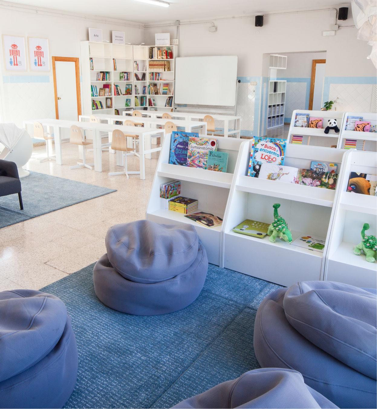 Biblioteca escolar amb zona de lectura infantil amb pufs grisos, catifes blaves, prestatgeries amb llibres i joguines, i taules amb cadires al fons.