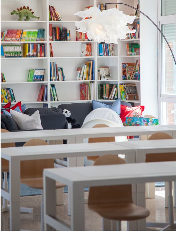 Biblioteca infantil amb prestatgeries plenes de llibres, sofàs amb coixins i peluixos, llum de peu blanca i taules amb cadires de fusta clara.