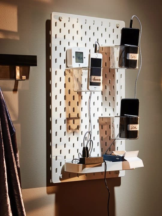 Biela perforovana nastenka IKEA SKÅDIS s priehľadnými úložnými nádobami na nabíjanie mobilných telefónov, praktické riešenie na organizáciu káblov a elektroniky v domácnosti.