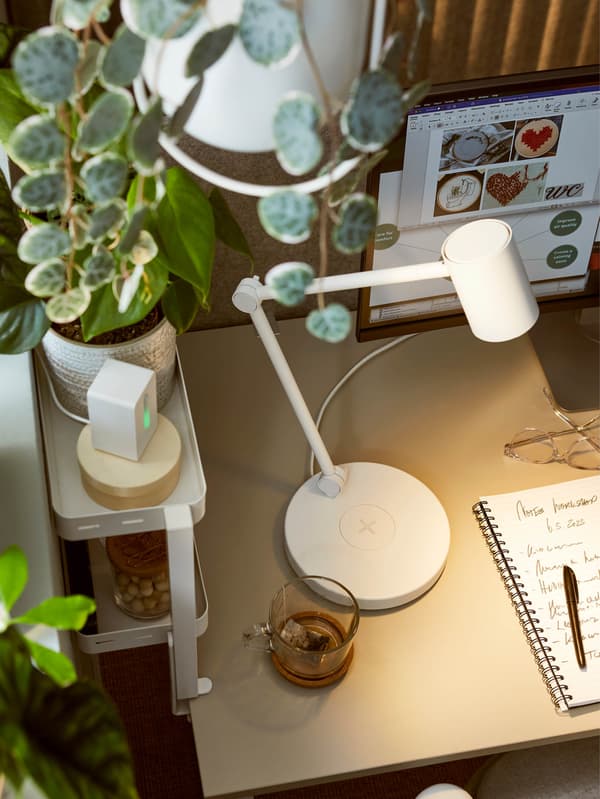 Biela polica na stôl VATTENKAR je pripevnená na stole, na ktorom stojí biela pracovná lampa NYMÅNE.