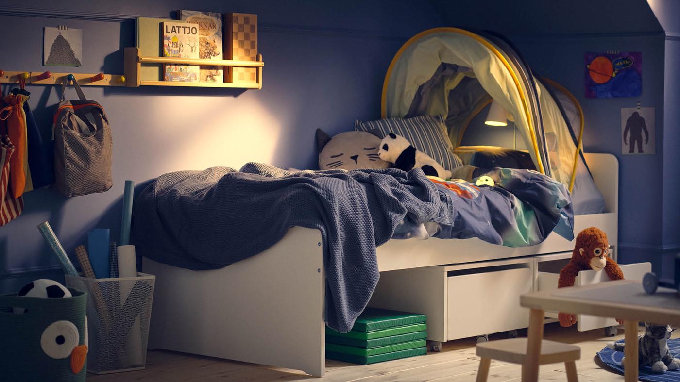 Biela posteľ SLÄKT s baldachýnom ELDFLUGA, úložným dielom s kolieskami SLÄKT a nástennou poličkou FLISAT s knihami.