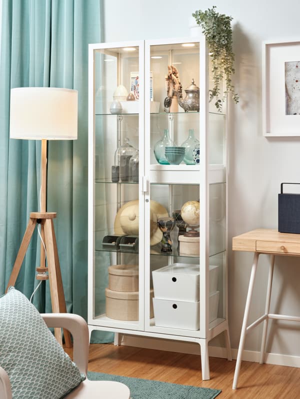 Biela zasklená vitrína MILSBO plná drobností, za ňou biela stena. Vľavo od vitríny je svetlotyrkysový záves, pred ním stojacia lampa LAUTERS. V popredí je stolička s tyrkysovým vankúšom, na podlahe tyrkysový koberec.