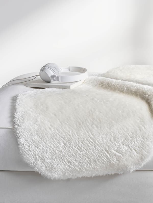 Biely koberec v tvare ovčej kožušiny TOFTLUND je prehodený cez bielu posteľ, na ňom leží biela kniha a biele slúchadlá.