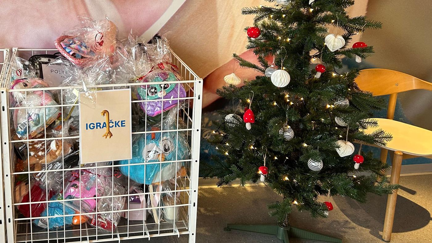 Bijeli metalni koš s oznakom „IGRAČKE“ ispunjen plišanim igračkama zamotanim u prozirnu foliju stoji uz ukrašeno božićno drvce s bijelim i crvenim kuglicama te svjetlima. U pozadini su drvene stolice.