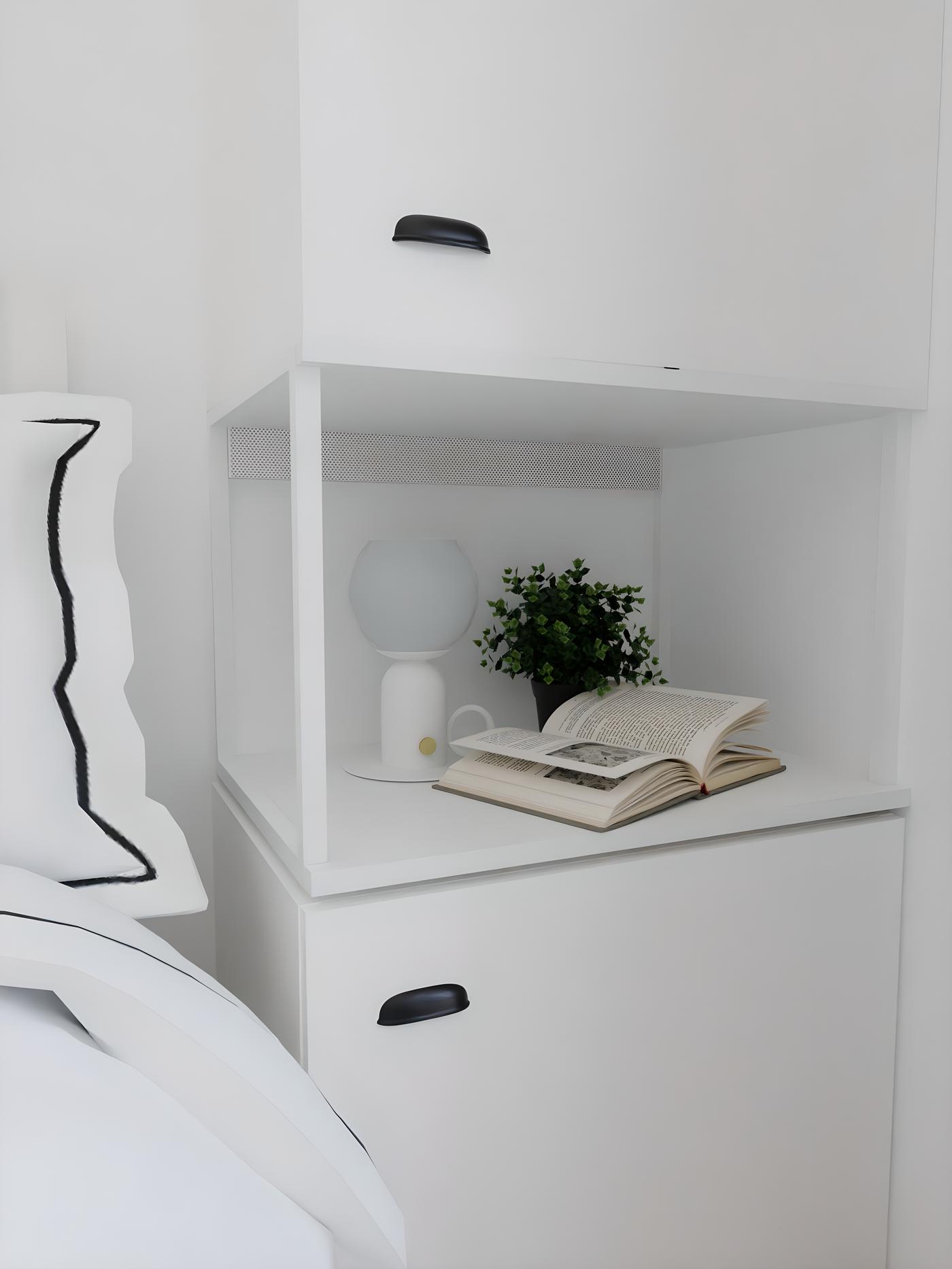 Bijeli noćni ormarić s otvorenom knjigom, biljkom u posudi i lampom na srednjoj polici.