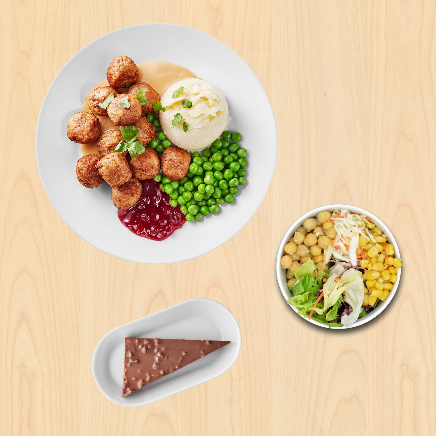 Bijeli tanjur s mesnim okruglicama, pire krumpirom, zelenim graškom i umakom, uz žlicu brusnica; pokraj zdjelica sa salatom od slanutka, kukuruza i povrća te tanjurić s komadom čokoladne torte.