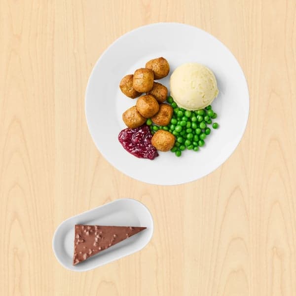Bijeli tanjur s ribljim okruglicama, pire krumpirom, zelenim graškom i žlicom brusnica na svijetloj drvenoj podlozi; ispod je mali tanjur s trokutastim komadom čokoladne torte.