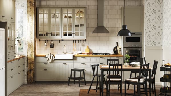 Bílá kuchyně provence v tradičním stylu s béžovými skříňkami, bílými obklady, dřevěnými podlahami, květinovými tapetami a černou jídelní soupravou.