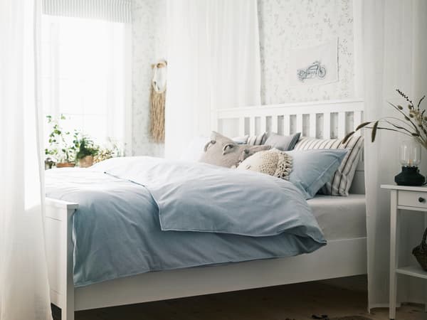 Bílá postel HEMNES ve světlé místnosti obklopená průhlednými bílými závěsy SILVERLÖNN s polštáři a modrým ložním prádlem.