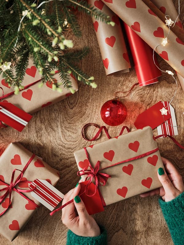 Bild tagen uppifrån på julklappar som slås in i brunt presentpapper med röda hjärtan på. Nertill i bilden syns en persons händer som håller i ett av paketen.
