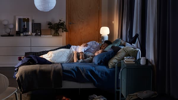Bilik tidur dengan rangka katil upholsteri SLATTUM, dua lampu dan sarung kuilt berwarna biru gelap, serta pasangan yang sedang tidur di atas katil.