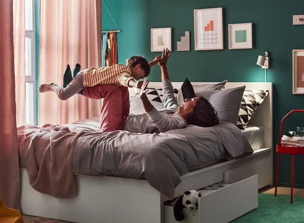 Bilik tidur dengan seorang ayah berbaring telentang sambil mengangkat tangan dan kakinya di atas katil, sedang bermain kapal terbang dengan anak lelakinya.