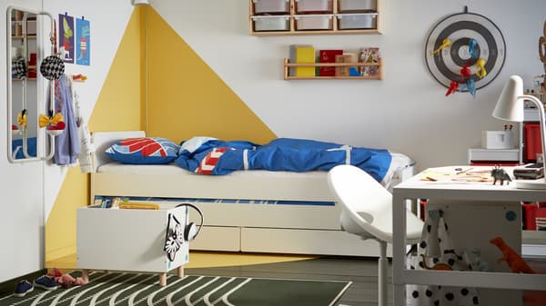 Bilik tidur kanak-kanak dengan dinding grafik berwarna putih dan kuning, katil berwarna putih dengan storan di bawahnya, linen katil berwarna biru dan merah.