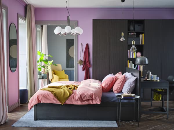 Bilik tidur yang berwarna-warni dengan dinding berwarna ungu lembut, linen katil berwarna coklat merah lembut, alas berwarna kuning di samping almari pakaian dan rangka katil berwarna hitam coklat.