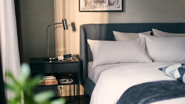 Bilik tidur yang disinari cahaya matahari dengan meja sisi katil berwarna hitam yang mempunyai buku dan lampu di sebelah katil TUFJORD berwarna biru dengan linen katil berwarna putih.