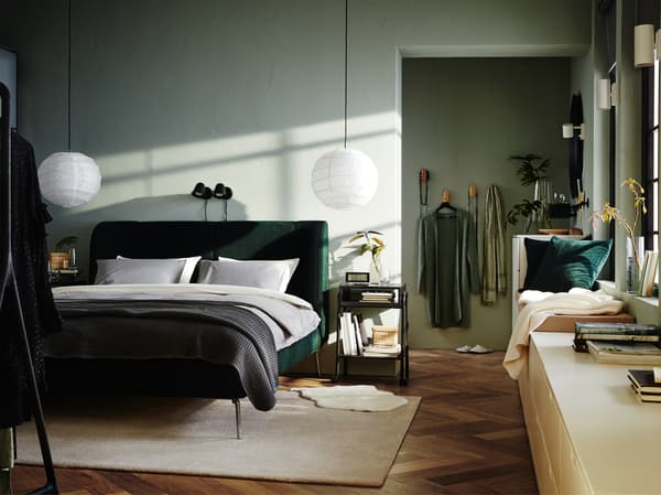 Bilik tidur yang mempunyai rangka katil upholsteri berwarna hijau, dua lampu dinding berwarna hitam, ambal berwarna putih pudar dan almari berlaci berwarna putih.