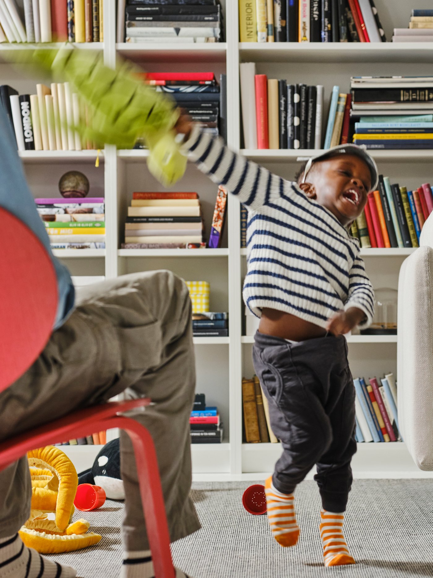 BILLY bokhyllor fyllda med böcker står mot en vägg bakom ett barn i mörka byxor och randig tröja som svingar en grön mjukleksak.