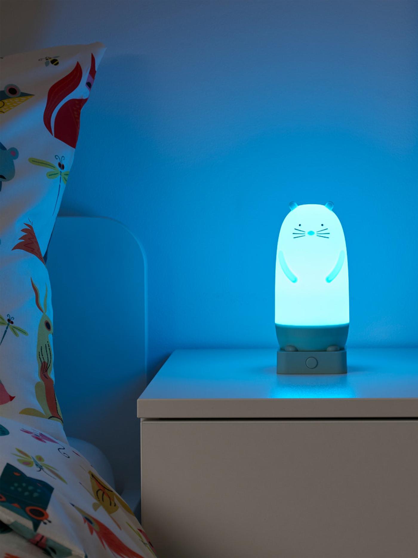 Bílý noční stolek s rozsvícenou noční lampičkou ve tvaru zvířátka, svítící na modro.